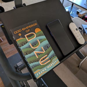 以下が含まれることがあります： 黒いトレッドミルのコンソールには、フランク・ハーバート著「デューンの神皇帝」のコピー、黒いスマートフォン、白いリモコンが置かれています。本の表紙は、オレンジと緑の色合いの風景デザインが特徴です。アイテムは黒い表面にあります。