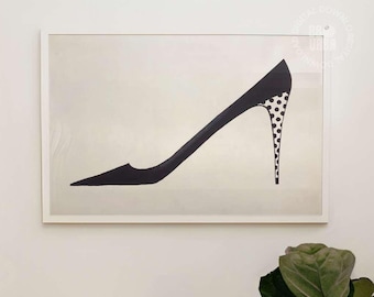 May include: A framed wall art print featuring a black stiletto shoe silhouette with a polka dot pattern on the heel. The artwork is displayed above a white toilet and a potted fiddle leaf fig plant in a woven basket within a bathroom.