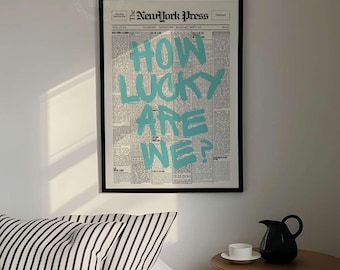 May include: A framed wall art print featuring a vintage newspaper page with the text New York Press and the large teal graphic text HOW LUCKY ARE WE? displayed above a bed with black and white striped bedding and a small wooden side table.