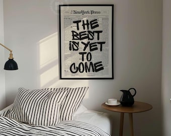 May include: A framed wall art print featuring the text New York Press and the phrase THE BEST IS YET TO COME in bold black lettering over a newspaper-style background. The print is displayed on a white wall above a bed with striped bedding.