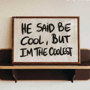 Op de afbeelding: Een rechthoekige houten lijst met een witte print met de zwarte tekst HE SAID BE COOL, BUT I'M THE COOLEST. De ingelijste print staat op een donkerbruine houten plank met afgeronde hoeken, voorzien van de tekst Digital Download in de hoek.