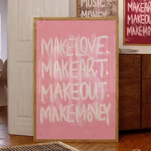 May include: A rectangular wall art print in a wooden frame featuring a pink background with white hand-painted style text that reads: MAKE LOVE. MAKE ART. MAKE OUT. MAKE MONEY. The print is displayed in a room setting with other framed wall art.