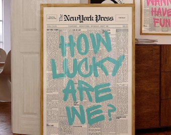 May include: A framed wall art print featuring a vintage newspaper layout titled The NewYork Press with the large, bold, light blue text HOW LUCKY ARE WE? overlaid on the center. The print is displayed in a light wood frame on a wooden floor.