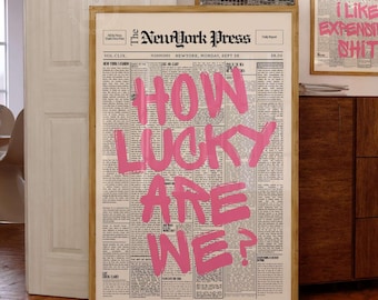 May include: A framed wall art print featuring a vintage newspaper layout titled The New York Press. The text HOW LUCKY ARE WE? is printed in large, bold, bright pink letters over the columns of black text on a light beige background.