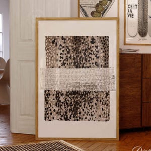 May include: Large framed artwork with a neutral color palette. The artwork features a textured design with a mix of spotted and woven patterns in shades of beige, gray, and brown. Additional framed art pieces and furniture are visible in the background.