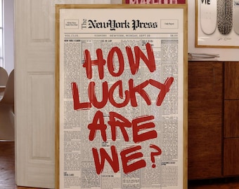 May include: A framed wall art print featuring a vintage newspaper layout titled The NewYork Press. The design displays the bold red hand-painted text HOW LUCKY ARE WE? centered over columns of small black printed news articles and text.