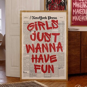 May include: A framed vertical wall art print featuring a vintage newspaper layout titled The NewYork Press with the bold red text GIRLS JUST WANNA HAVE FUN overlaid across the center. The print is displayed in a light wood frame on a floor.
