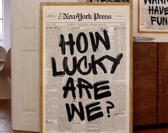 May include: A framed wall art print featuring a vintage newspaper layout titled The New York Press with the bold black hand-lettered text HOW LUCKY ARE WE? centered on the page. The design mimics a classic broadsheet newspaper with columns.