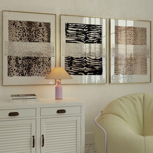 May include: Three framed wall art prints featuring animal print patterns in black, white, and brown, each overlaid with a horizontal textured band. A small table lamp with a pink base and a pleated shade sits on a white cabinet with louvered doors.