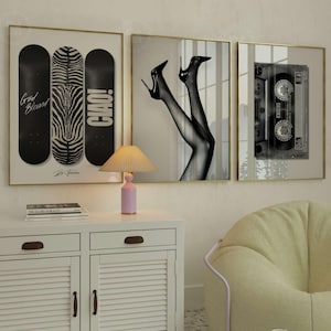 Könnte beinhalten: Drei gerahmte Wandbilder mit einer schwarz-weißen Skateboard-Grafik mit Text, einem Paar Beine in High Heels und einer Kassette. Darunter steht eine rosa Tischlampe mit plissiertem Schirm auf einem weißen Schrank mit Lamellentüren.