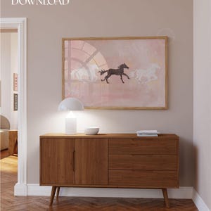 K&ouml;nnte beinhalten: Ein rechteckiger gerahmter Wandkunstdruck mit einer zentralen schwarzen Pferdesilhouette, flankiert von zwei wei&szlig;en Pferdesilhouetten vor einem zartrosa Hintergrund mit einem kreisf&ouml;rmigen Bogendesign. Der Text Digital DOWNLOAD steht oben links.