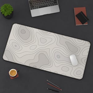 May include: A large rectangular desk mat with a topographic map design in shades of gray and black. A white computer mouse rests on the mat, alongside a laptop, a small potted plant, and stationery.