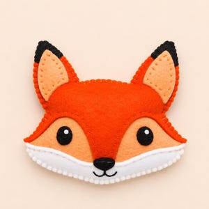 Könnte beinhalten: Ein Fuchs-Applikation aus Filz mit leuchtend orangefarbenem Fell, hellpfirsichfarbenen Akzenten an Ohren und Gesicht sowie einem weißen Kinn. Der Fuchs hat aufgestickte schwarze Augen, eine schwarze Nase und schwarze Ohrspitzen.