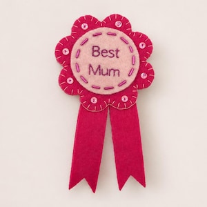 Peut inclure: Un badge en feutre rose vif avec l'inscription "Best Mum" au centre. Le badge a la forme d'une fleur avec un ruban et est orné de coutures et de petites perles. Un cadeau attentionné.
