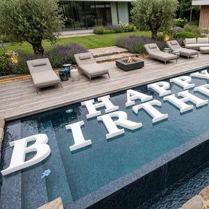 Puede incluir: Una piscina con las letras blancas "HAPPY BIRTHDAY" flotando en el agua. La piscina está rodeada por una terraza de madera con tumbonas y una pequeña hoguera. El paisaje incluye árboles y flores.