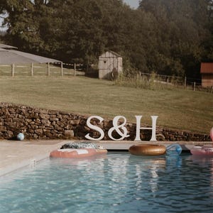 May include: Large white freestanding letters S, &, and H positioned on a stone wall edge beside a swimming pool. The pool contains several colorful inflatable pool floats, including a gold ring and a pink float, under a clear sky.