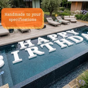 Könnte beinhalten: Weiße, schwimmende Buchstaben, die "HAPPY BIRTHDAY" in einem Swimmingpool buchstabieren. Der Pool ist von einer Holzterrasse und Gartenmöbeln umgeben. Der Text "Handmade to your specifications" befindet sich in einem orangefarbenen Rechteck.