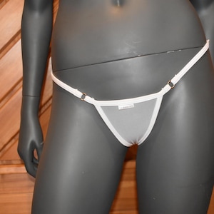 Peut inclure: String blanc avec fines bretelles et petits accents métalliques dorés. Le tissu transparent révèle un panneau avant triangulaire. Le vêtement est présenté sur un mannequin sur fond de bois.