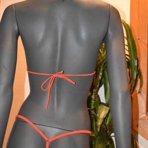 May include: An orange bikini set on a mannequin. The bikini top ties at the neck and back, and the bottom is a thong style. The mannequin is gray, and the background includes a plant and a wooden frame.