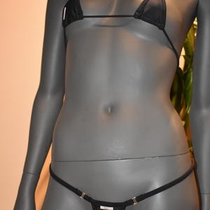 May include: Black bikini set on a mannequin. The top is a triangle style with thin straps. The bottom is a thong style with gold-colored accents. The fabric appears to be a smooth, solid color.