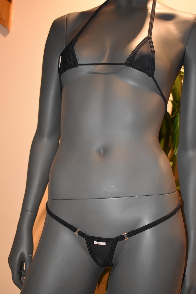 May include: Black bikini set on a mannequin. The top is a triangle style with thin straps. The bottom is a thong style with gold-colored accents. The fabric appears to be a smooth, solid color.