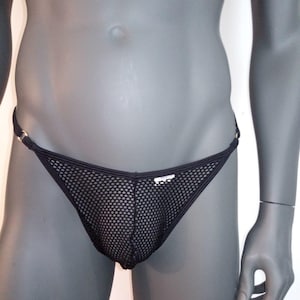 May include: Black mesh thong underwear with a low-rise waist. The underwear features a small, rectangular tag on the front and gold-colored metal ring accents on the sides. The fabric has a net-like pattern.