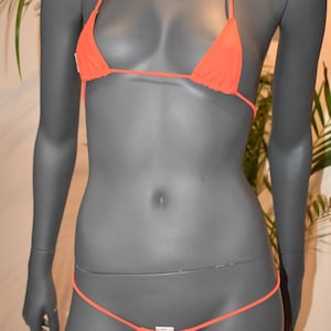 May include: Orange bikini set on a mannequin. The triangle top has thin straps and the bottom is a thong style. The swimwear is a vibrant shade of orange, perfect for beach or pool wear.