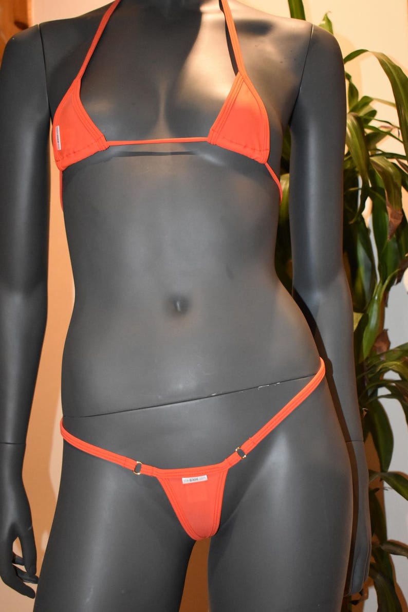 May include: An orange bikini set displayed on a mannequin. The set includes a triangle top with halter straps and a thong-style bottom. The bikini is made of a smooth fabric and features small metal ring details.