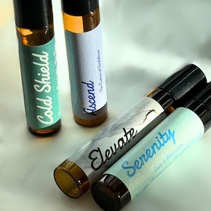 May include: Four glass roller bottles with blue and white labels. The labels have the words "Cold Shield", "Ascend", "Elevate", and "Serenity" on them.