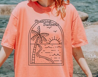 May include: Coral-colored t-shirt featuring a black line art design of a palm tree, ocean, and sunset, with the words "Chasing Sunsets" above. The shirt has short sleeves and a relaxed fit.