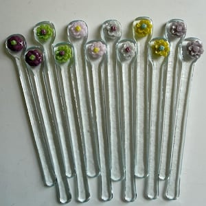 May include: A collection of clear glass stir sticks, each topped with a colorful, detailed flower. The flowers feature shades of purple, green, pink, and yellow. The sticks are slender and appear to be approximately 6 inches long.