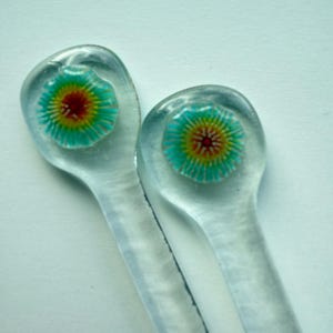 May include: Two clear glass hair pins with a colorful floral design. The flower has a turquoise outer ring, yellow and red center. The pins have a rounded top and a long, slender stem.