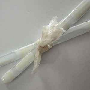 May include: Two clear glass tubes with a white, creamy substance inside, tied together with a cream-colored, frayed ribbon. The tubes are angled across each other against a white background.