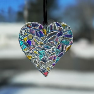May include: A mosaic heart ornament with a black string for hanging. The heart is composed of iridescent glass pieces in shades of silver, blue, pink, yellow, and green. The background is blurred, suggesting an outdoor setting.