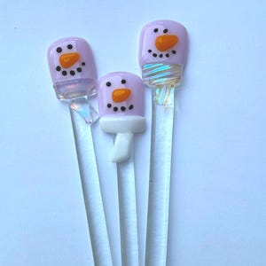 May include: Three whimsical snowman-themed glass stir sticks. Each stick features a lavender head with an orange carrot nose and black dot eyes and mouth. One snowman has a white scarf. The clear glass sticks are perfect for holiday drinks.