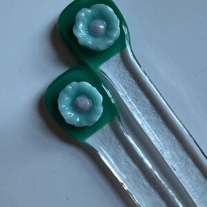 May include: Two glass stir sticks with a green square top featuring a light blue flower with a pink center. The sticks are clear with a white stripe down the center. The background is a light, neutral color.