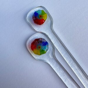 May include: Two clear glass stirring sticks with colorful glass flower designs at the top. The flowers have a rainbow pattern with red, yellow, blue, green, and orange colors.