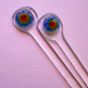 May include: Two clear glass stir sticks with colorful, spiral-shaped designs on the ends. The spirals feature concentric rings of red, yellow, green, and blue. The sticks are on a light purple background.