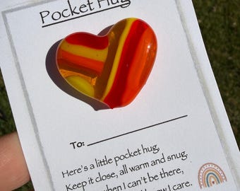 May include: A colorful glass heart-shaped pocket hug with yellow, orange, and red stripes, resting on a white card. The card reads "Pocket Hug" and includes a message about keeping it close. The Etsy shop URL is also visible.