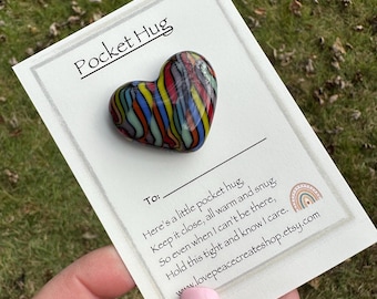 May include: A small, colorful heart-shaped glass ornament with a striped pattern. The ornament is on a white card with the text "Pocket Hug" and a message about keeping the hug close.