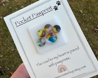 May include: A white card with the text "Pocket Pawprint" and a small white square with five colorful glass marbles. The text below the square reads "The road to my heart is paved with pawprints." and "www.lovepeacecreateshop.etsy.com".