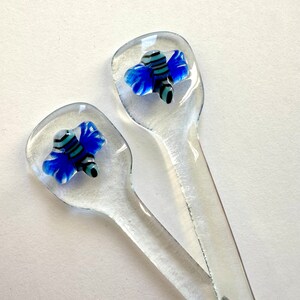 May include: Two clear glass spoons with a blue and black bee design on the spoon head. The bee has blue wings and a black and white striped body. The spoons are on a white background.
