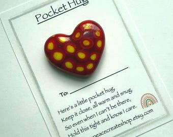 May include: A red heart-shaped pocket hug with yellow polka dots, resting on a white card. The card reads "Pocket Hug" and includes a message about warmth and care. The Etsy shop URL is also visible.