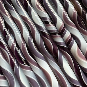 May include: Close-up of several twisted glass rods with a swirl pattern. The rods are primarily white, lavender, and deep purple, creating a striped effect. The rods are arranged diagonally, filling the frame.