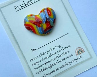 May include: A multi-coloured glass heart-shaped pocket hug rests on a card. The heart has swirls of red, yellow, blue, and white. The card is printed with the words "Pocket Hug" and a message about comfort and care, with a website address.
