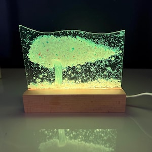 May include: A glowing glass tree sculpture with a wooden base. The tree is illuminated with green light, and the glass has a textured, speckled appearance. The base is rectangular and made of light-colored wood. A white cord extends from the base.