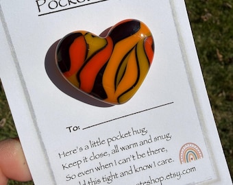 May include: A heart-shaped pocket hug with a swirling pattern of orange, yellow, and black. The card reads "Pocket Hug" and includes a message about keeping it close. The Etsy shop URL is also visible.