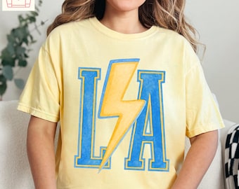 Puede incluir: Camiseta amarillo claro con las letras "LA" en azul, contorneadas en azul más oscuro, y un rayo amarillo en el centro. La camiseta es Comfort Colors 1717 Butter.