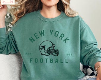 Könnte beinhalten: Ein hellgrüner Pullover mit einer schwarzen Grafik eines Footballhelms, über dem ein Flugzeug fliegt. Der Text "NEW YORK" befindet sich über dem Helm und "EST. 1963 FOOTBALL" unter dem Helm.