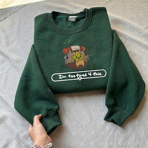 Puede incluir: Sudadera verde oscuro con cuello redondo y mangas largas. La parte delantera presenta un gráfico bordado de un personaje de dibujos animados con una mochila. Debajo, el texto "I'm too tired 4 this." La sudadera está doblada sobre una superficie clara.